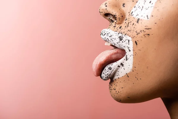 Foto recortada de chica afroamericana con salpicaduras de pintura en la cara mostrando la lengua aislada en rosa - foto de stock