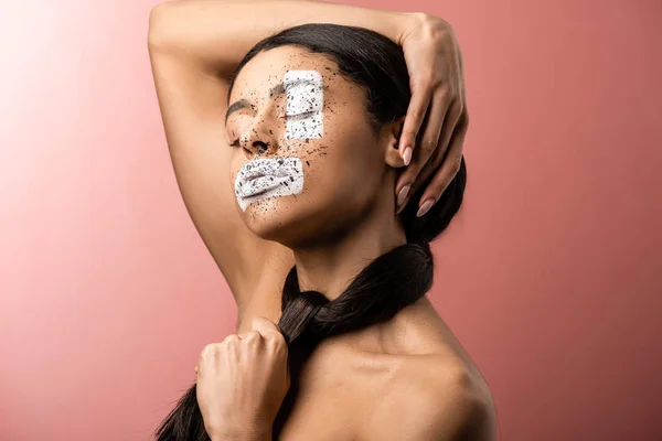 Atractiva chica afroamericana sensual con la cara pintada tocando la cabeza aislada en rosa - foto de stock