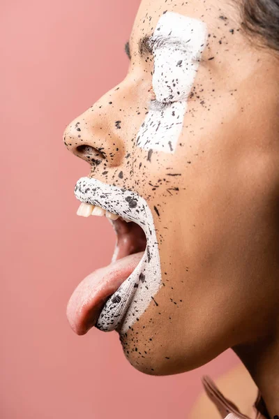 Vue latérale de la fille afro-américaine avec des éclaboussures de peinture sur le visage montrant la langue isolée sur rose — Photo de stock