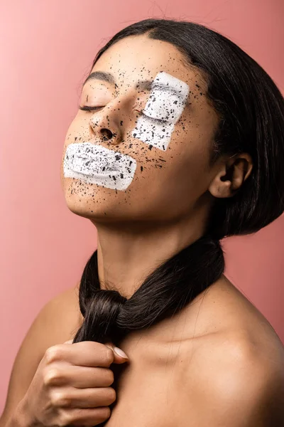 Hermosa mujer afroamericana con salpicaduras de pintura en la cara y el pelo alrededor del cuello aislado en rosa - foto de stock