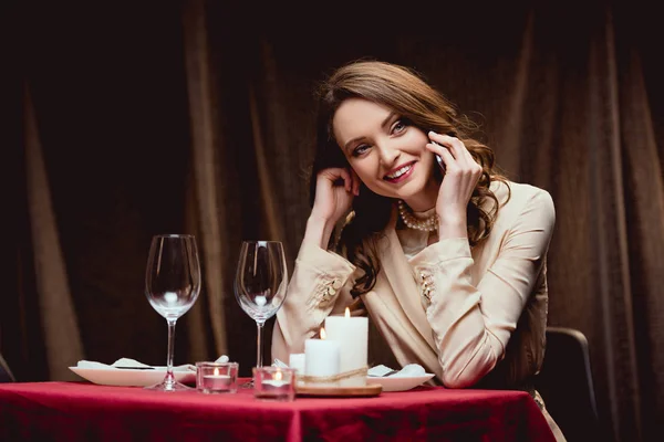 Hermosa mujer sonriente sentada en la mesa y hablando en el teléfono inteligente en el restaurante - foto de stock