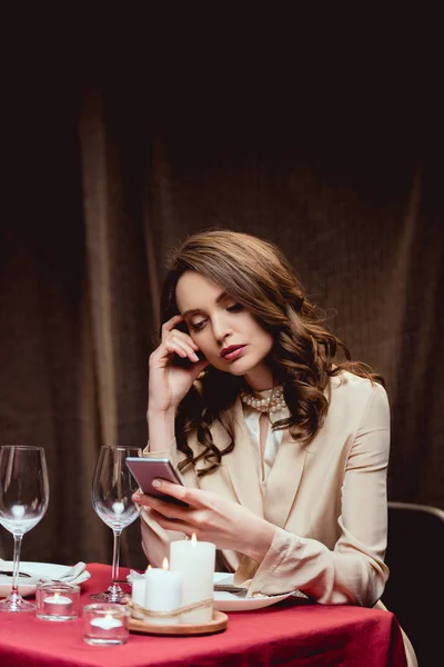 Hermosa mujer pensativa sentada en la mesa y el uso de teléfono inteligente en el restaurante - foto de stock