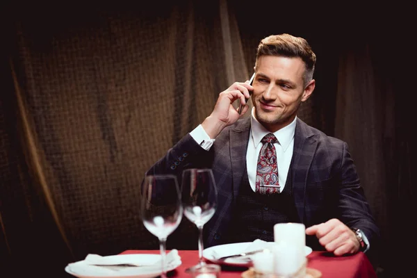 Hombre sonriente guapo en traje sentado en la mesa y hablando en el teléfono inteligente en el restaurante — Stock Photo