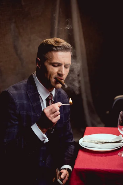 Handsome man in suit sitting at table and lighting up cigar in restaurant — Stock Photo