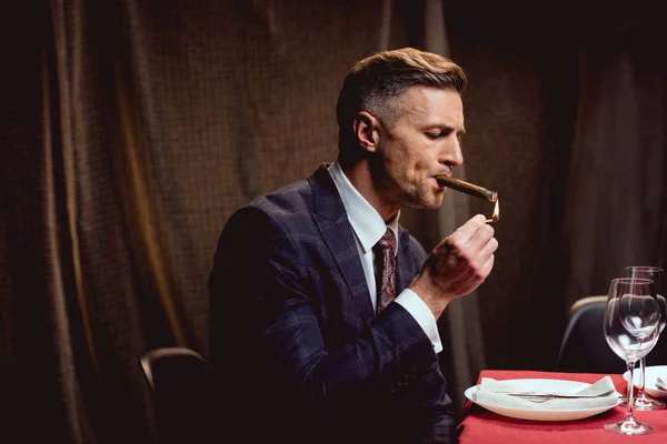 Schöner Mann im Anzug sitzt am Tisch und zündet sich Zigarre im Restaurant an — Stockfoto