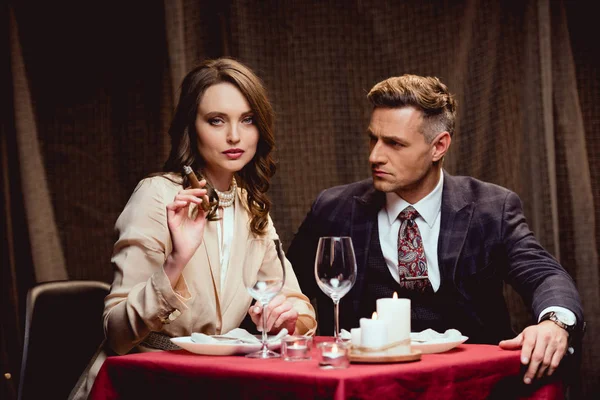 Belle femme assise à table et fumant du cigare tout en ayant un dîner romantique avec un bel homme au restaurant — Photo de stock