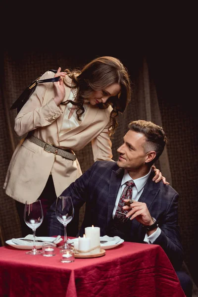 Mujer abrazando sonriente hombre con cigarro durante la cena romántica en el restaurante - foto de stock