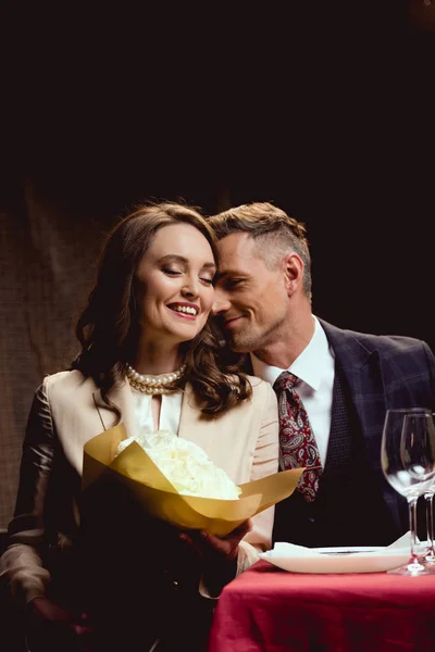 Красивая счастливая пара, сидящая за столом с цветочным букетом во время романтического свидания в ресторане — стоковое фото