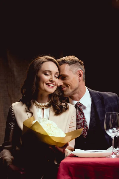 Beau couple heureux assis à table avec bouquet de fleurs pendant la date romantique au restaurant — Photo de stock