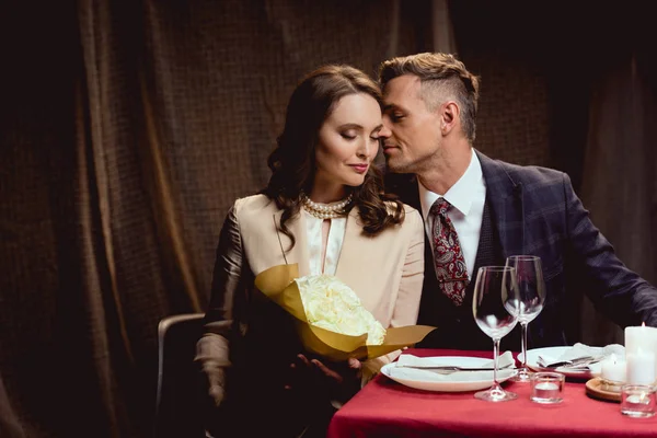 Hermosa pareja sentada en la mesa con ramo de flores durante la cita romántica en el restaurante - foto de stock