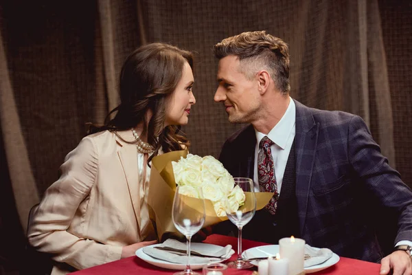 Hermosa pareja sentada en la mesa con ramo de flores y mirándose durante una cita romántica en el restaurante - foto de stock