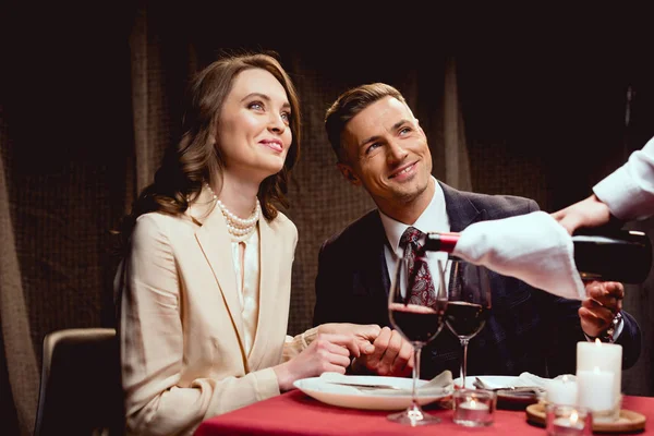 Camarero vertiendo vino tinto mientras sonríe pareja teniendo cita romántica en el restaurante - foto de stock