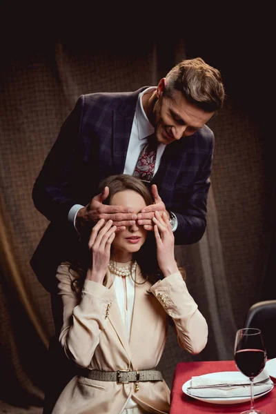 Homem sorridente cobrindo olhos de mulher com mãos de surpresa durante a data romântica no restaurante — Fotografia de Stock