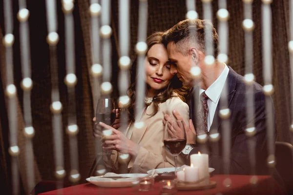 Hombre abrazando suavemente a la mujer durante la cita romántica en el restaurante con luces bokeh en primer plano - foto de stock