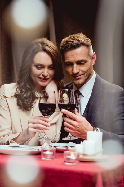 Sonriente pareja tintineo vasos de vino tinto durante la cita romántica en el restaurante con luces bokeh en primer plano - foto de stock