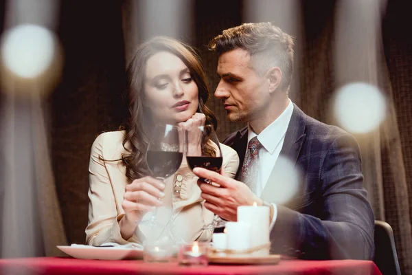 Pareja tintineo vasos de vino tinto durante la cita romántica en el restaurante con luces bokeh en primer plano - foto de stock