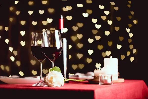 Table setting with glasses of red wine, rose and heart shaped bokeh on background — Stock Photo