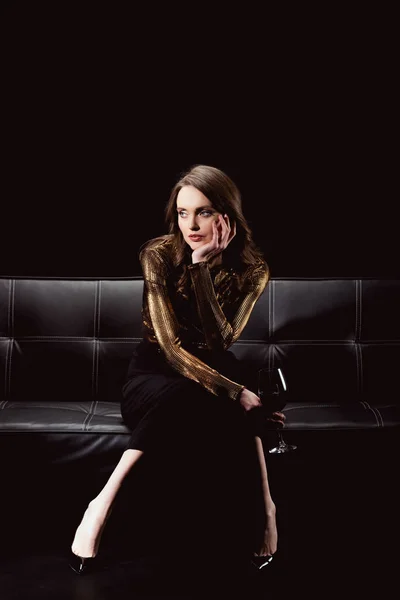 Beautiful glamorous woman sitting on couch with glass of red wine and propping chin isolated on black — Stock Photo