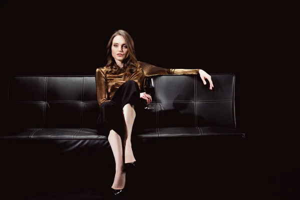 Bella donna glamour seduta sul divano con bicchiere di vino rosso isolato su nero — Foto stock