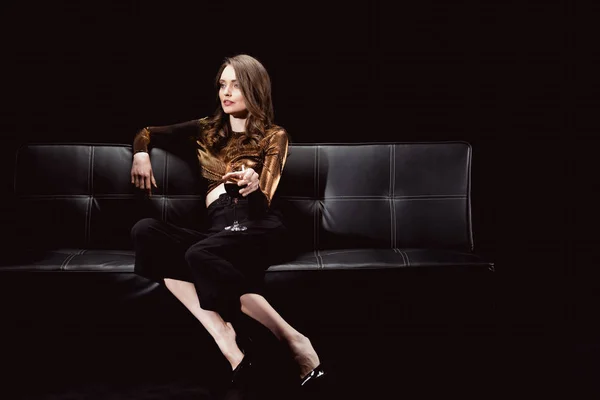 Beautiful glamorous woman sitting on couch and posing with glass of red wine isolated on black — Stock Photo