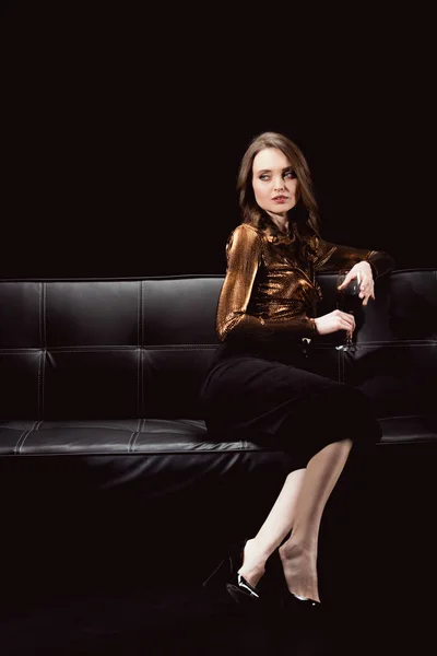 Belle femme glamour assis sur le canapé avec un verre de vin rouge et regardant loin isolé sur noir — Photo de stock