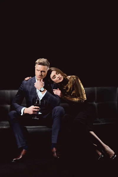 Beautiful woman sitting on couch and embracing man with glass of red wine isolated on black — Stock Photo