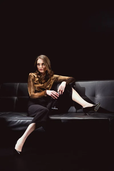 Hermosa mujer glamorosa sentada en el sofá y posando con copa de vino tinto aislado en negro - foto de stock