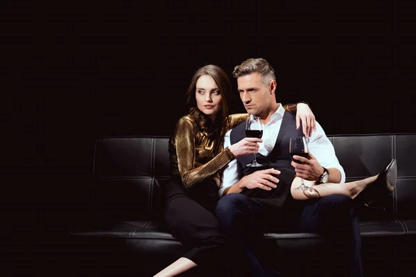 Beautiful couple sitting on couch with glasses of red wine isolated on black — Stock Photo