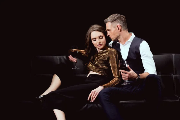 Beautiful romantic couple sitting on couch with glasses of red wine isolated on black — Stock Photo