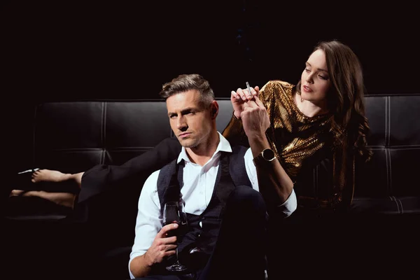 Beautiful woman lying on couch with cigarette while handsome man holding glass of red wine isolated on black — Stock Photo