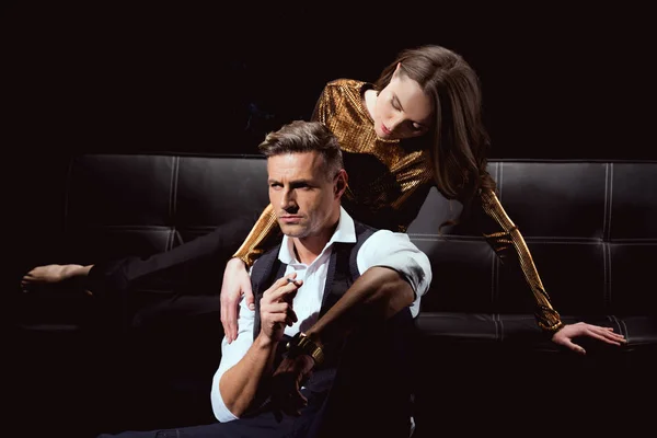Hermosa mujer acostada en el sofá y abrazando hombre guapo con cigarrillo aislado en negro - foto de stock