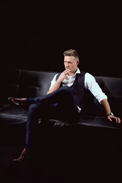 Серьезный красивый мужчина в формальной одежде сидит на диване с сигаретой изолированы на черный — стоковое фото