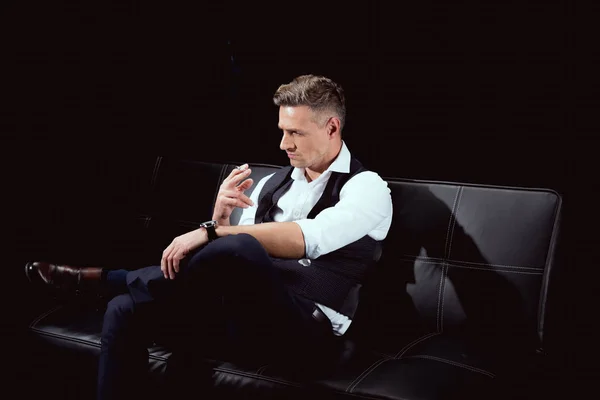 Handsome man in formal wear sitting on couch with cigarette isolated on black — Stock Photo