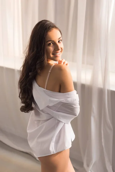 Hermosa mujer sonriente en camisa blanca mirando a la cámara y posando en casa por la mañana - foto de stock