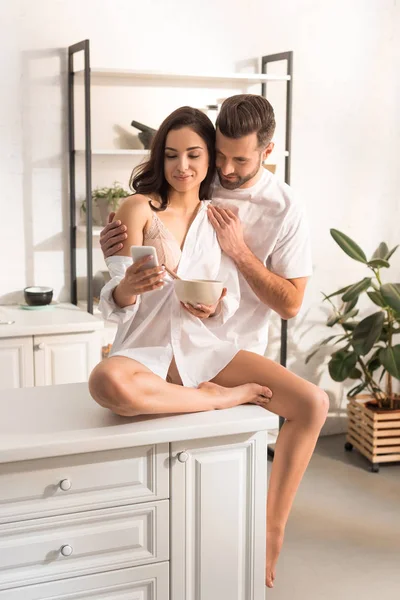 Mann umarmt schöne Frau beim Frühstück in Küche mit Smartphone — Stockfoto