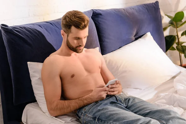 Красивый мужчина без рубашки лежит в постели и использует смартфон дома утром — стоковое фото