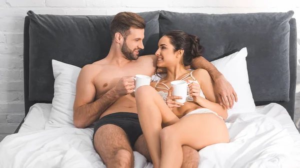 Beautiful sexy couple lying with coffee cups in bed, smiling and looking at each other — Stock Photo