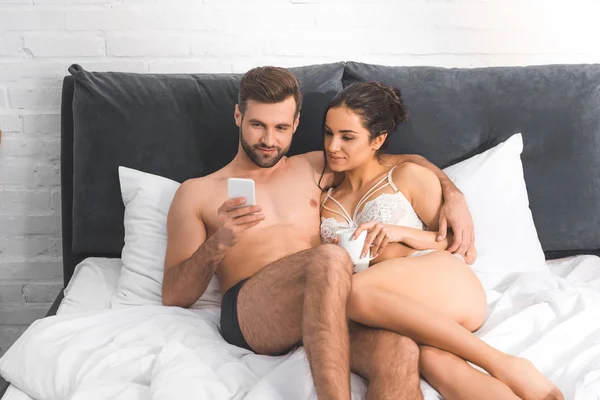 Hombre guapo abrazando a mujer hermosa y el uso de teléfono inteligente en la cama - foto de stock