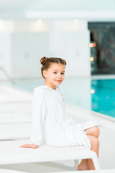 Fröhliches Kind sitzt auf Liegestuhl im Bademantel neben Schwimmbad — Stockfoto