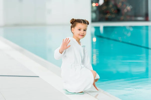 Mignon enfant assis dans peignoir ner piscine et agitant la main en disant bonjour — Photo de stock