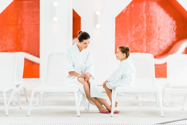 Happy mother looking at daughter in bathrobe while sitting on deck chair — Stock Photo