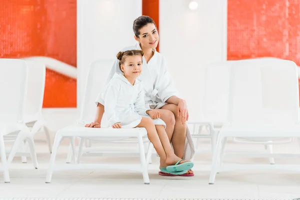 Happy mother sitting with daughter on deck chairs and smiling while looking at camera — Stock Photo