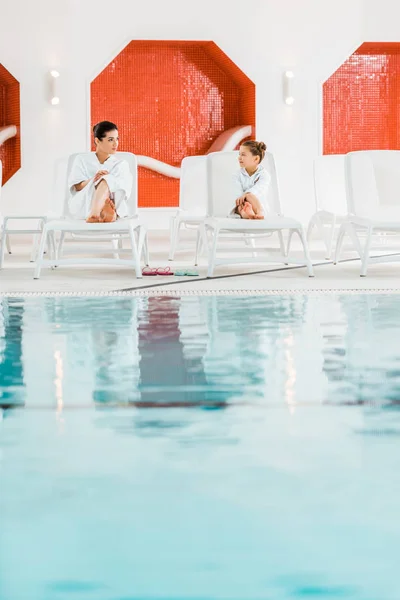 Fröhliche Mutter liegt mit süßer Tochter im Bademantel auf Liegestühlen — Stockfoto