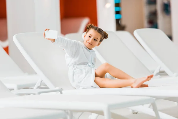 Cheerful child in bathrobe taking selfie while lying on deck chair — Stock Photo