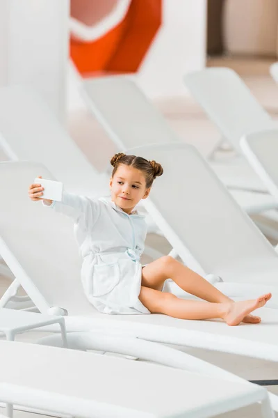 Cute child in bathrobe taking selfie while lying on deck chair — Stock Photo