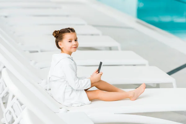 Cute child in white bathrobe holding smartphone near swimming pool — Stock Photo