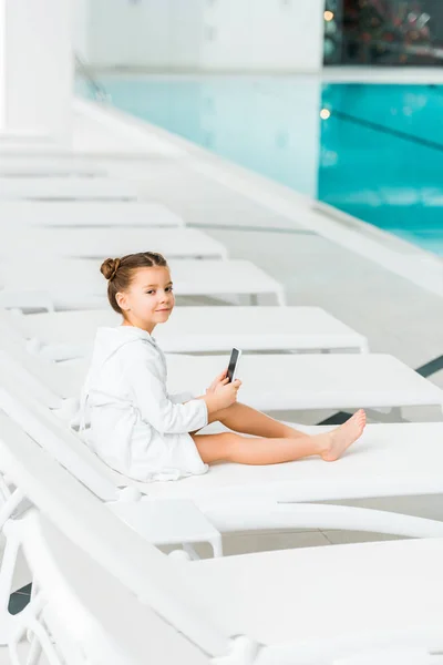 Selektiver Fokus des niedlichen Kindes im weißen Bademantel, das Smartphone in der Nähe des Swimmingpools hält — Stockfoto