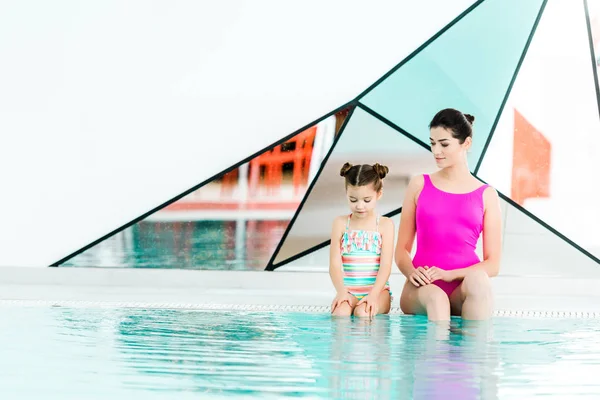 Attraente madre seduta con carina figlia in costumi da bagno vicino alla piscina — Foto stock