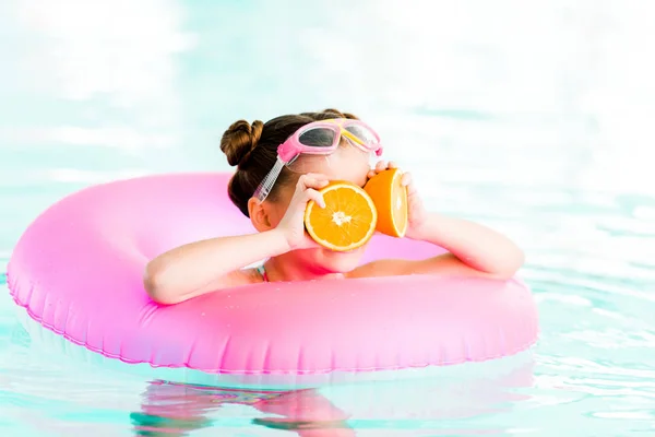 Niño sosteniendo media naranja cerca de los ojos mientras nada con anillo inflable en la piscina - foto de stock