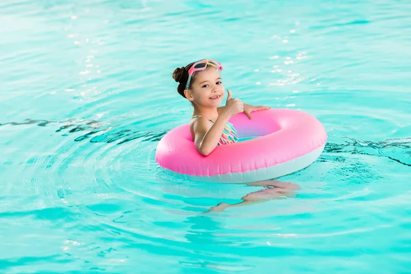 Criança feliz nadando com anel inflável e mostrando sinal do polegar para cima na piscina — Fotografia de Stock